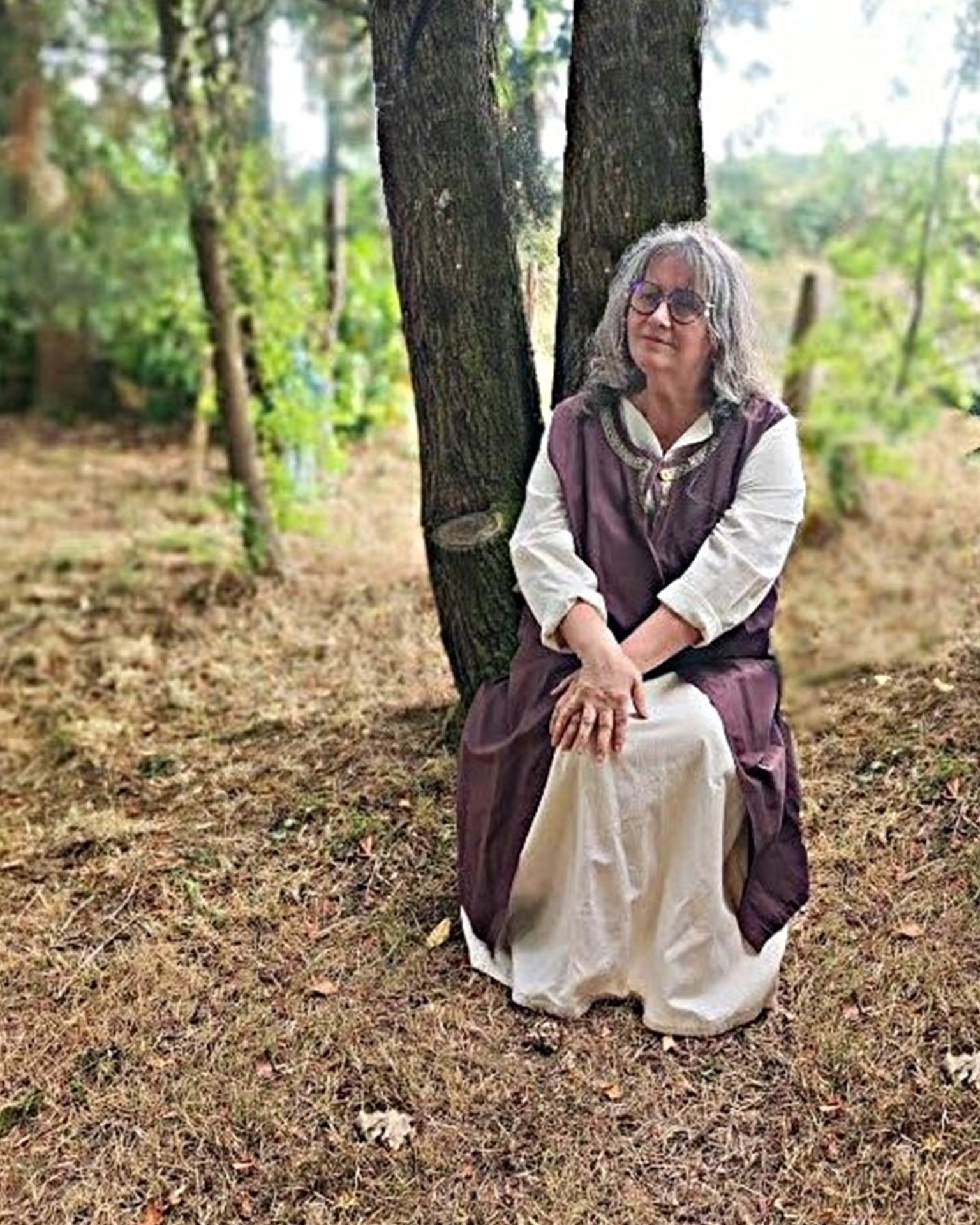 Femme assise près d’un arbre, portant une robe blanche et un gilet marron, en forêt.