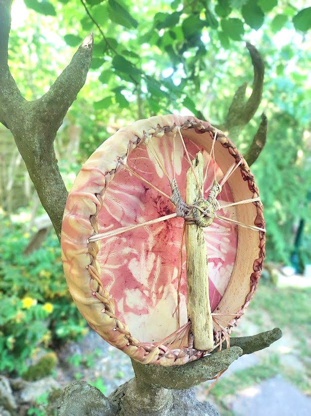 Tambour circulaire en peau, décoré de bois, cordes, et anneau, posé sur une sculpture de cerf dans un jardin vert.