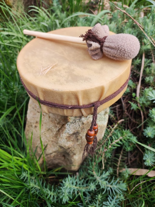 Petit tambour en bois posé sur une souche, recouvert d’une peau de cerf beige, dans l’herbe verte.