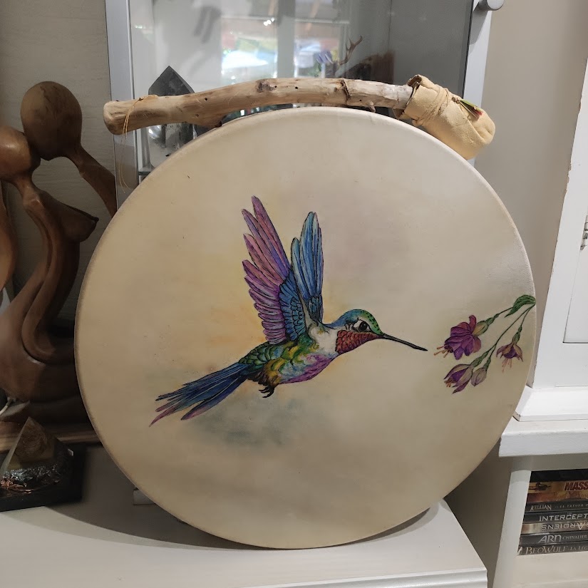 Tambour rond beige avec peinture d’un oiseau multicolore en vol et fleurs violettes, posé en vitrine.